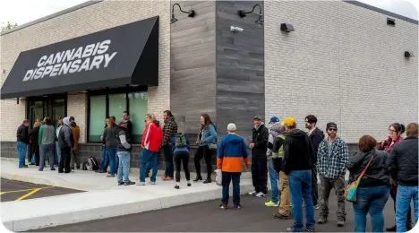 A line of people waiting outside a modern building with a black awning labeled "Cannabis Dispensary."