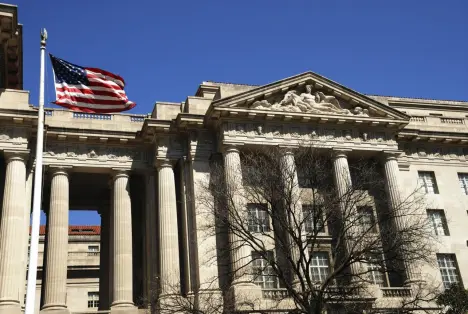 The facade of a classic stone government building featuring large columns with an American flag flying in the foreground.