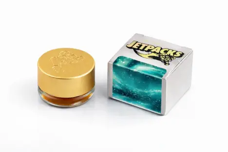 A small glass concentrate jar with a gold lid sitting next to a white Jetpacks retail box with a turquoise design.