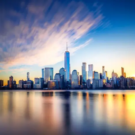A wide-angle view of the Lower Manhattan skyline and One World Trade Center reflecting on the water during a vibrant sunset.