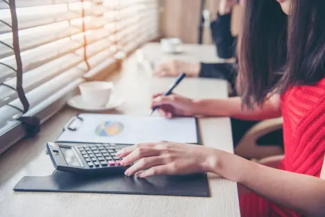 A person at a desk using a calculator and writing on a financial document with a laptop and a cup of coffee nearby.