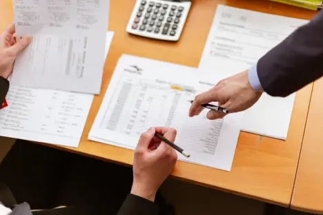 Two people sitting at a wooden table reviewing financial spreadsheets and tax documents with pens in hand.